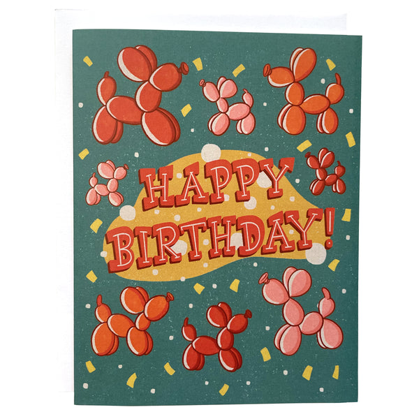 A turquoise birthday card features red, orange and pink balloon dogs, yellow confetti, and the words "Happy Birthday!" in midcentury-inspired hand lettering. The card sits against a white envelope on a white background.