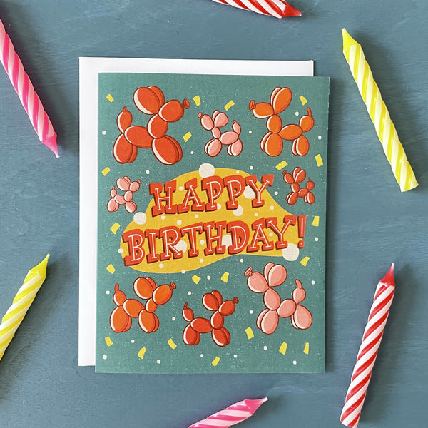 A turquoise birthday card features red, orange and pink balloon dogs, yellow confetti, and the words "Happy Birthday!" in midcentury-inspired hand lettering. The card is on a blue wooden background surrounded by striped birthday candles.