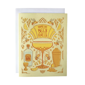 A yellow art-deco inspired greeting card is covered with illustrations of bees, honey and cocktail accessories, along with a crystal coupe filled with a Bee's Knees cocktail. A marquee on the card reads "You're the Bee's Knees" in dark orange letters. The card sits against a white envelope on a white background.