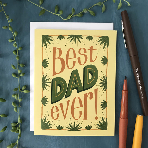 A pale yellow card reads "Best DAD ever!" in brown and green hand lettering. The letters are framed in a brown rectangle out of which sprout green tropical plants. The card rests on a blue wooden background along with brown, orange and yellow pens and some strands from a string-of-pearls plant.