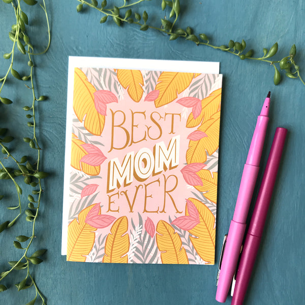 A pink card covered in golden yellow, white and grey tropical leaves with the lettered phrase "Best Mom Ever" rests on a blue background beside 2 pink pens. Buds from a string of pearls plant surround it.⁠