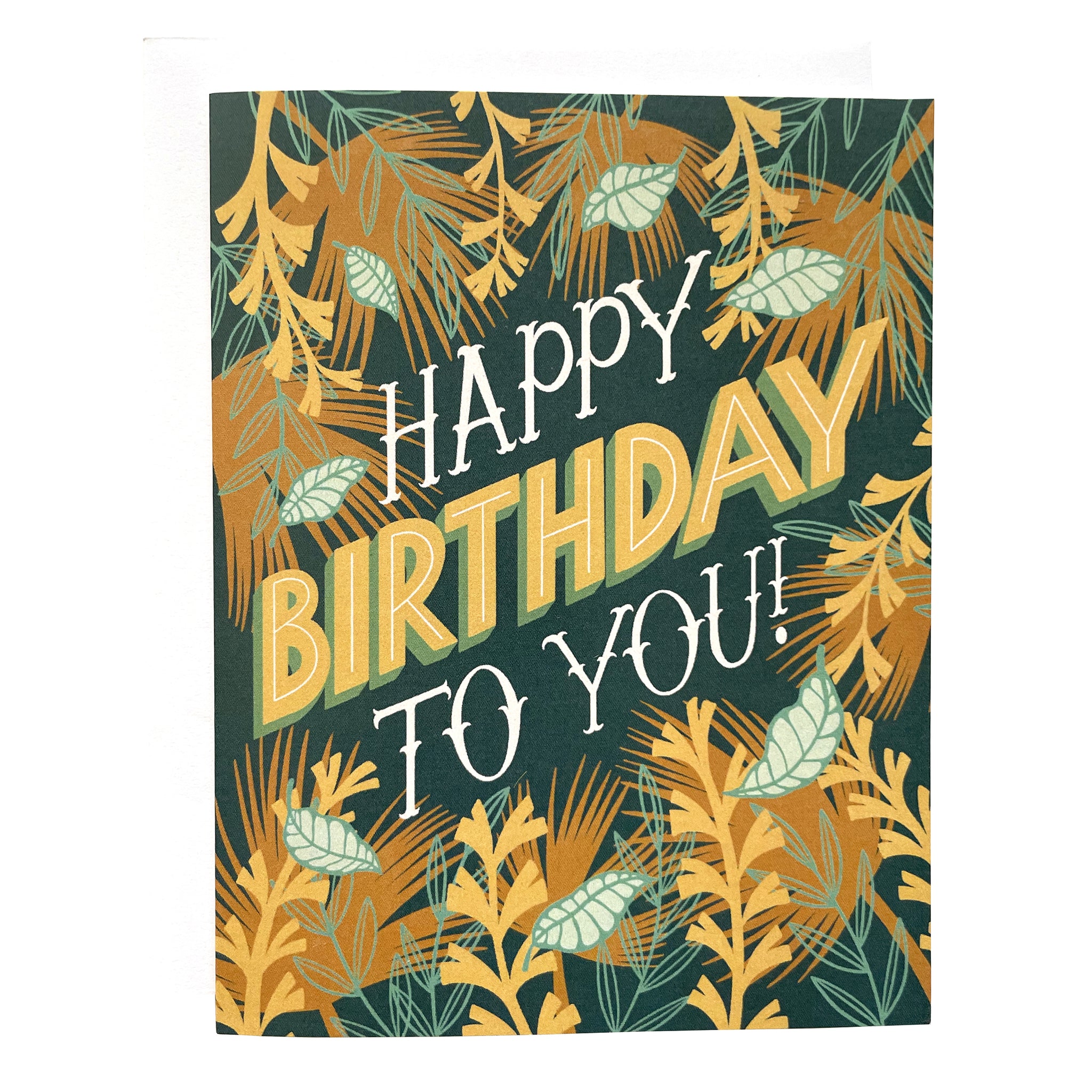 A dark blue greeting card features the hand-lettered words "Happy Birthday to you!". The letters are framed by illustrations of tropical leaves and vines. The card sits against a white envelope on a white background.
