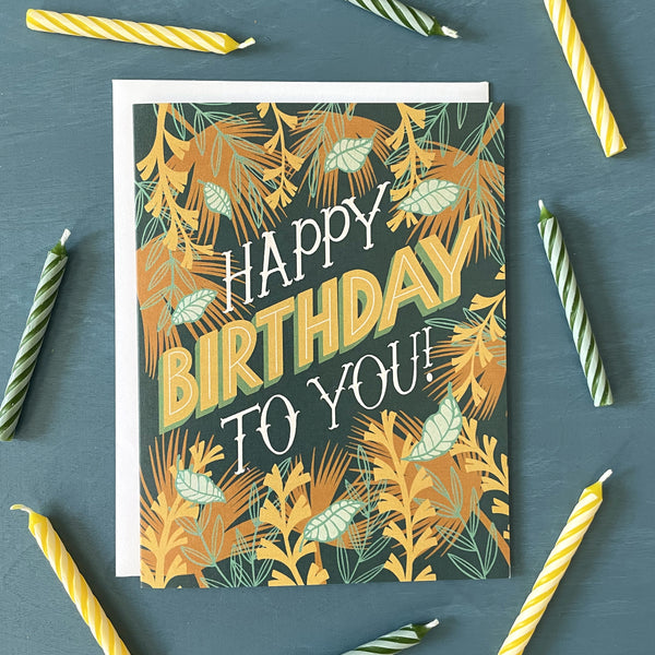 A dark blue greeting card features the hand-lettered words "Happy Birthday to you!". The letters are framed by illustrations of tropical leaves and vines. The card is surrounded by striped birthday candles.