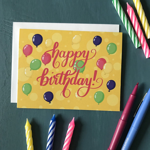 A yellow greeting card is surrounded by pens and striped birthday candles. On the card are the words "happy birthday," lettered in bright pink, and several brightly coloured balloons.