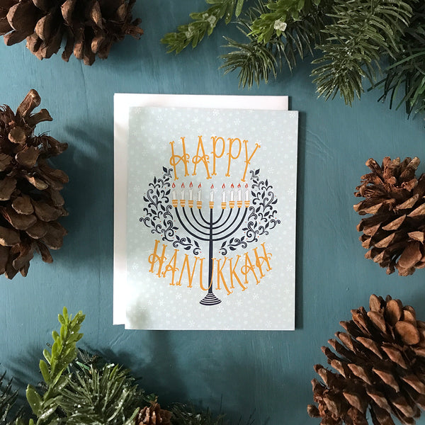 A pale blue snowflake-patterned greeting card is flanked by faux greenery and pinecones. The card reads Happy Hanukkah in yellow hand-lettering. In between the words is a navy menorah with lit candles flanked by navy flourishes. 