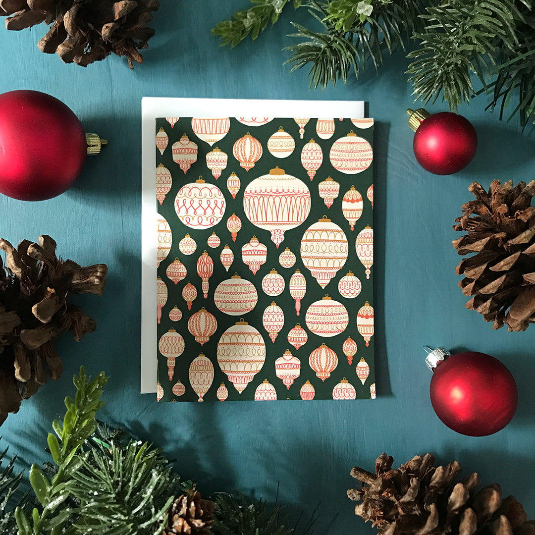 A green greeting card boasts a pattern of cream, red and gold tree ornaments. The card is surrounded by red Christmas ornaments, pinecones, and faux winter greenery on a blue wooden background.⁠