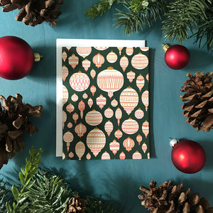 A green greeting card boasts a pattern of cream, red and gold tree ornaments. The card is surrounded by red Christmas ornaments, pinecones, and faux winter greenery on a blue wooden background.⁠
