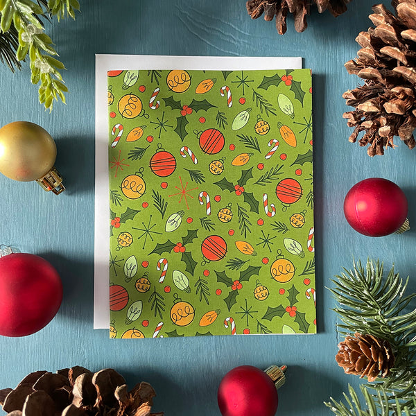 A green greeting card is patterned with midcentury-inspired ornaments, holly, candy canes and Christmas lights. The card is on a blue background and is flanked by ornaments, pinecones and faux greenery.