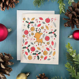 A blush pink card features a cute reindeer surrounded by an explosion of Christmas lights, ornaments, pine branches and holly. The card is surrounded by ornaments, pinecones and faux greenery.