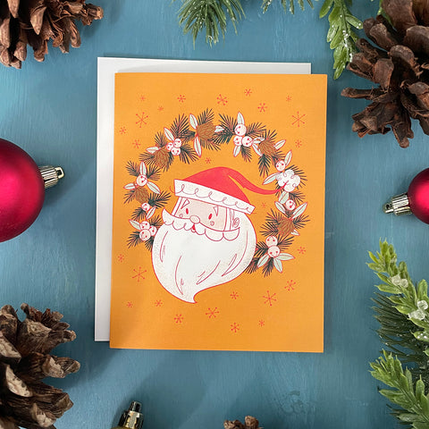 A yellow card shows a midcentury-style illustration of Santa's head in a wreath of jingle bells, pinecones and evergreen branches. The card is surrounded by ornaments, pinecones and faux greenery.