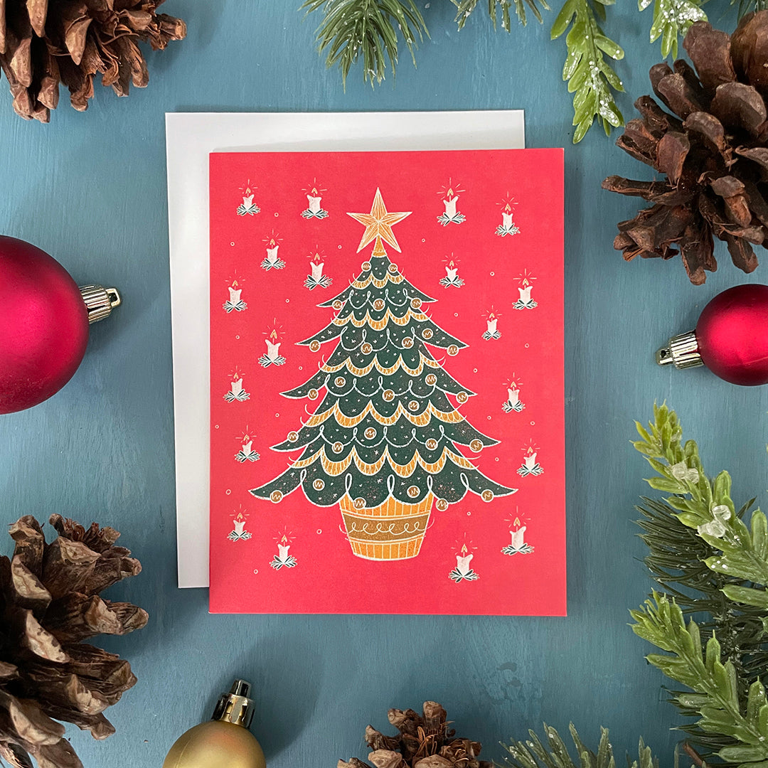 A red card features a midcentury-style bushy evergreen tree in a pot covered in baubles and garland, around which is a pattern of lit candles. The card is surrounded by ornaments, pinecones and faux greenery.