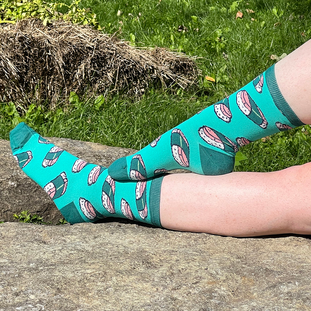 A pair of pale, freckled legs sports a turquoise pair of socks patterned with pink-and-green musubi-style sushi. One foot is outstretched on a rock while the other foot rests on top of that leg, all in front of a grassy background.