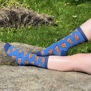 A pale, freckled pair of legs wears a pair of blue socks patterned with pepperoni pizza slices. The cuff, heel and toe of the socks are darker blue. The legs are on a rock against a grassy background.