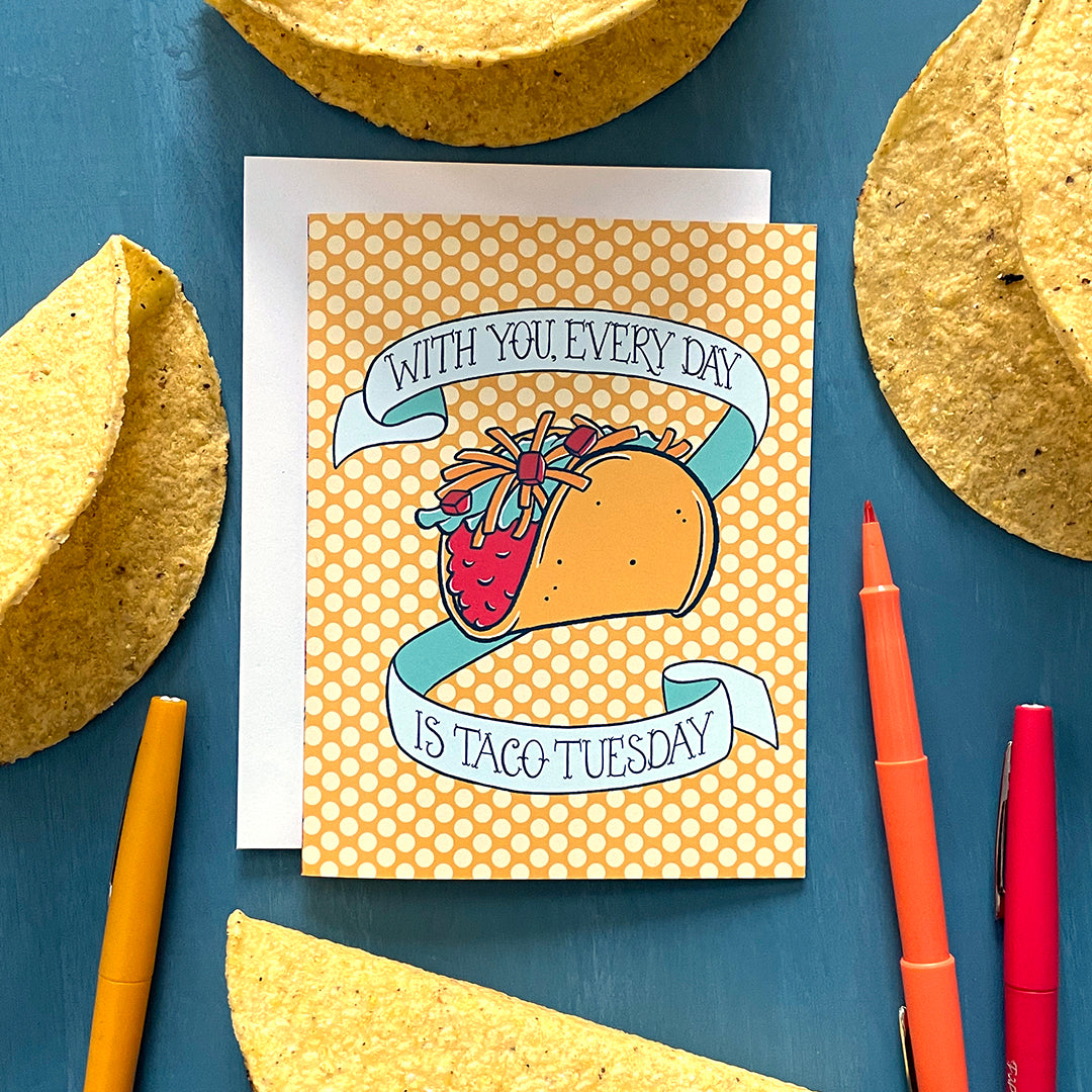A yellow polka-dot greeting card features an illustration of a hard-shell taco loaded with toppings and the words With You, Every Day is Taco Tuesday in hand-lettering. The card is on a white envelope and is surrounded by taco shells and red, yellow and orange pens on a blue background.