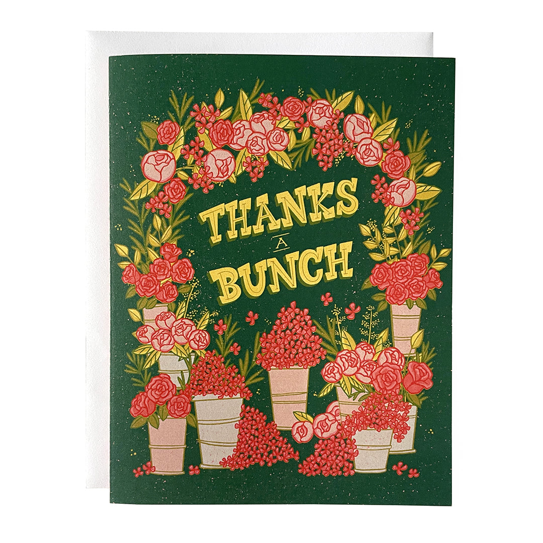 A dark green greeting card shows an arch covered in pink flowers with buckets of flowers at the bottom. It reads Thanks a Bunch in yellow letters. It is against a white envelope on a white background.