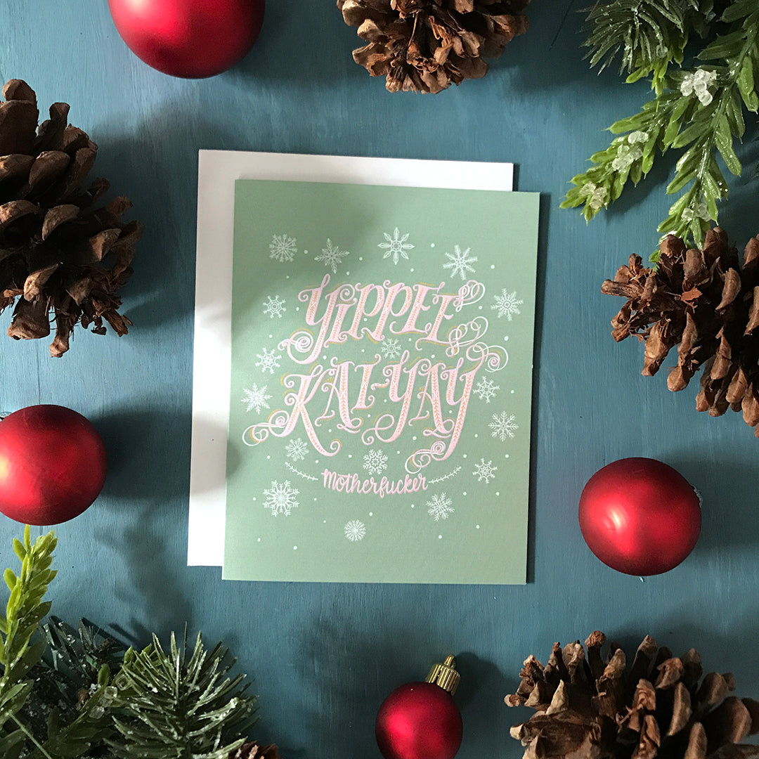 A light green card with pale mint snowflakes shows the words Yippee Kai-Yay, motherfucker in elaborate pink lettering. The card is flanked by pinecones, ornaments and faux greenery.