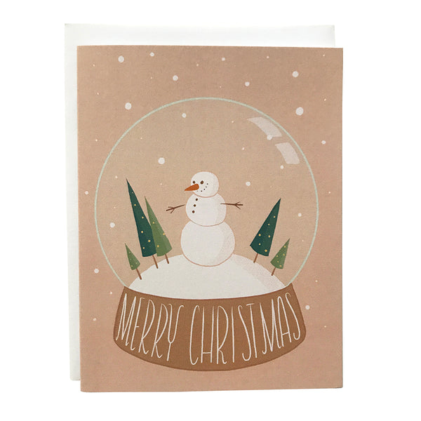 A pink Christmas card features an illustration of a snow globe with a cheerful snowman, triangular evergreens, and the words Merry Christmas lettered at its base. The card sits against a white envelope on a white background.
