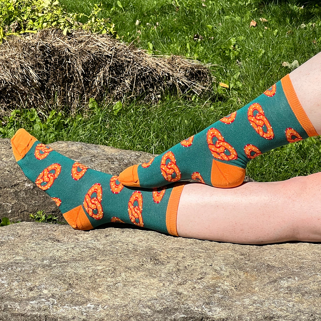 A pale, freckled pair of legs wears a pair of dark green socks patterned with orange salted pretzels and with orange heels, cuffs and toes.  The legs are against a rock in a grassy background.