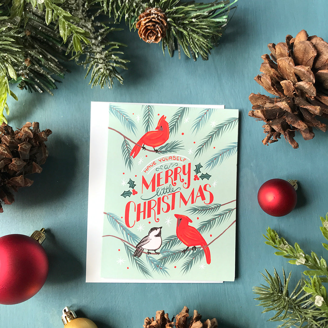 A greeting card reads Have Yourself a Merry Little Christmas lettered in red and green, surrounded by cardinals and a chickadee on a mint background. The card is surrounded by pinecones, Christmas ornaments and faux evergreen branches.⁠