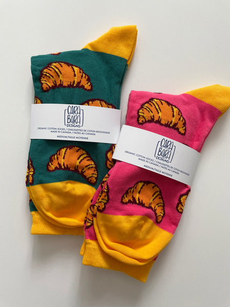 A pair of teal croissant-patterned socks and a pair of pink croissant-patterned socks are folded and labelled with belly bands with the Carabara Designs logo and the words organic cotton socks made in Canada, size medium, in English and French.
