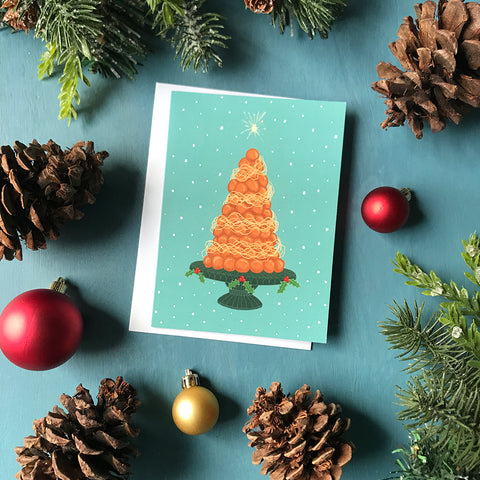 A light blue card shows a tall croquembouche sitting on a green, holly-laden pedestal platter. A sparkler is lit at the top of the dessert. The card is flanked by ornaments, pinecones and faux greenery.