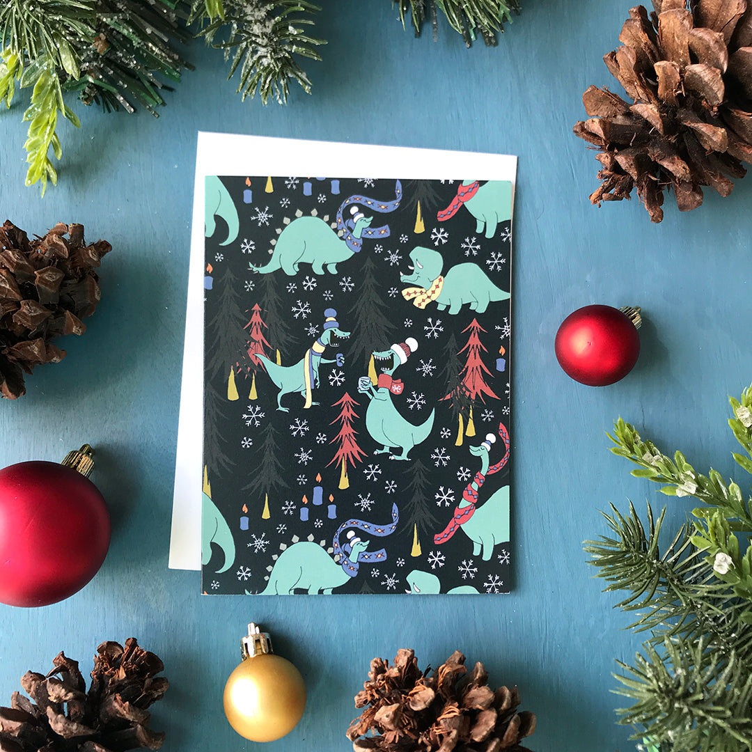 A greeting card is covered with a pattern of dinosaurs in toques and scarves with mugs of hot chocolate. Scattered among them are trees and candles. The card is on a blue background surrounded by red Christmas ornaments, pinecones and faux greenery.