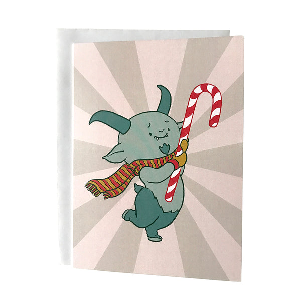 A pink card features a cute horned demon wearing mittens and a scarf and hugging a candy cane. The demon stands on two hooved legs. The card is on a white envelope on a white background.