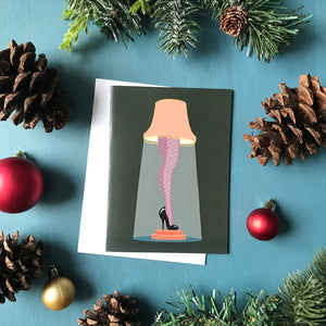 A dark green greeting card features an illustration of a lampshade atop a leg clad in fishnet stockings with a black high heel. The card is surrounded by pinecones, red Christmas ornaments and faux greenery.