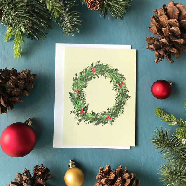 A pale green greeting card with an illustration of an evergreen wreath adorned by holly berries. The card is surrounded by pinecones, red Christmas ornaments, and faux greenery on a blue wooden background.