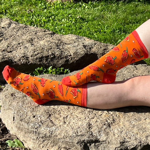 A pair of pale freckled bare legs wears orange socks patterned with red-and-yellow hot peppers with red cuffs, heels and toes. The photo was taken outdoors on a rock near grass.