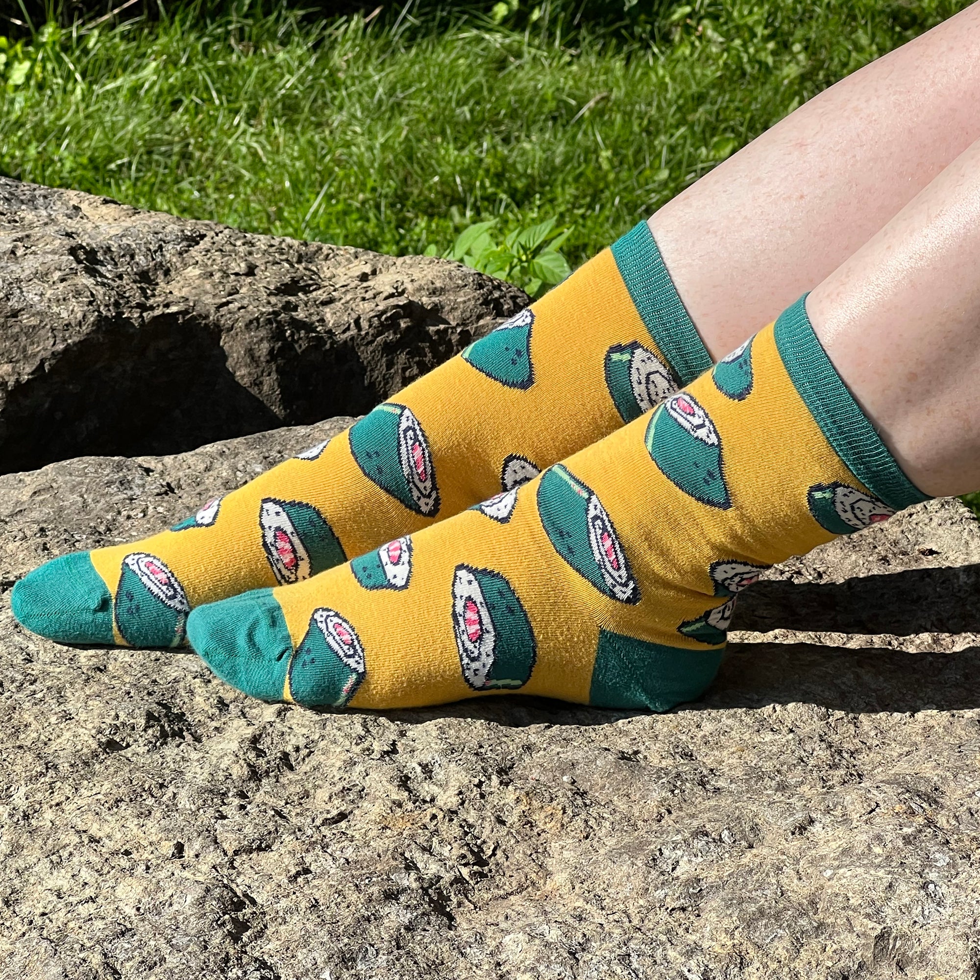 A pair of pale, freckled legs sports a yellow pair of socks patterned with seaweed-wrapped sushi rolls. One foot is outstretched on a rock while the other foot rests on top of that leg, all in front of a grassy background.